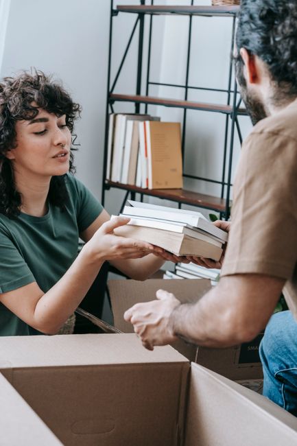 A woman with curly dark hair, wearing a green t-shirt, is inside a room holding a stack of books wrapped in plastic or paper, while a man with dark hair and a beard, dressed in a brown shirt, is reach