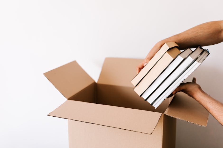 A woman with curly dark hair, wearing a green t-shirt, is inside a room holding a stack of books wrapped in plastic or paper, while a man with dark hair and a beard, dressed in a brown shirt, is reach