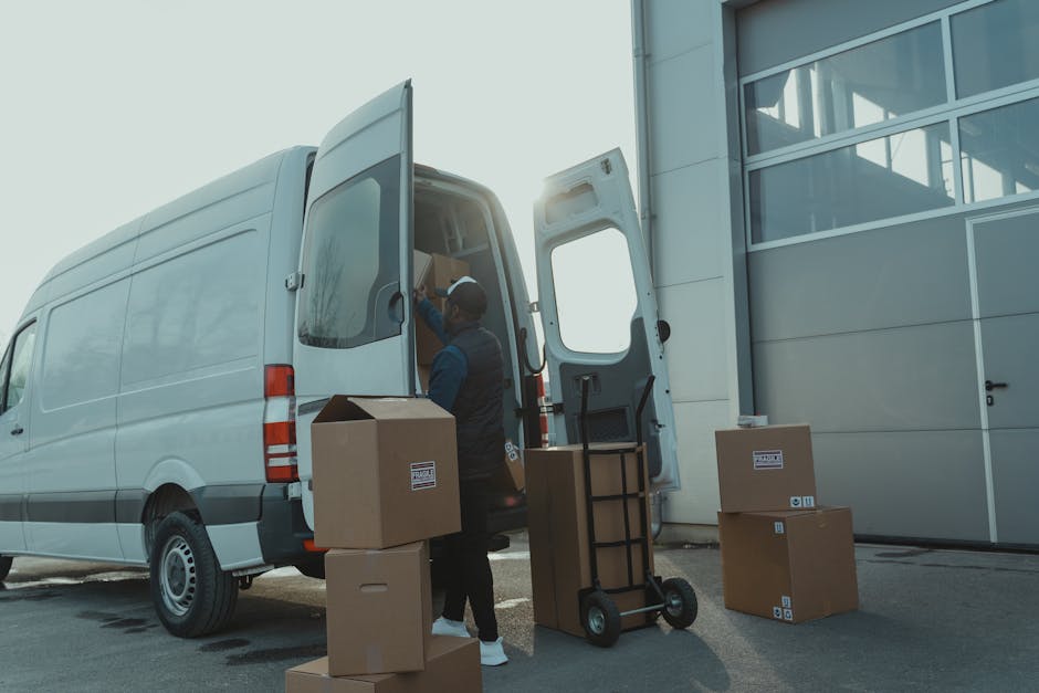 A man wearing a white T-shirt, jeans, and a black cap is outside a white van, which is parked on a residential street in Maida Vale. He is carrying three large cardboard boxes stacked vertically, with
