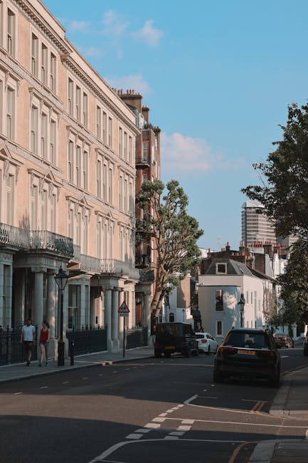A residential street scene showing a row of multi-storey Victorian-style houses with cream-colored facades, large windows, and decorative wrought iron balconies. The street is lined with mature trees, casting shadows on parked cars including a black hatchback and a white van, with another vehicle partially visible behind. On the pavement, a person is seen walking past a black lamp post, while another stands near the entrance of one property. The sky above is blue with scattered clouds, and there is a modern high-rise building visible in the background. In the context of home relocation, a moving team from Man With a Van Maida Vale could be involved in the loading process, using furniture blankets and packing materials to protect items during transport, with a van positioned nearby for furniture and box loading in preparation for a house move in Sutherland Avenue, Maida Vale.