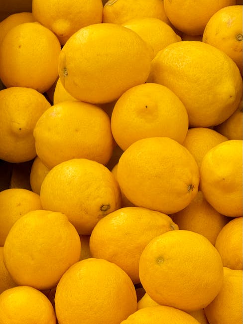 A close-up view of numerous bright yellow lemons piled together, with some showing small blemishes and slight variations in size, occupying the majority of the frame. The lemons appear fresh with a smooth, slightly textured skin. The image is taken indoors, with natural or diffused lighting illuminating the fruit evenly. This detailed photograph of lemons is relevant to house removals and packing processes, where fresh produce may need to be carefully transported during a home relocation or furniture transport, highlighting the importance of secure packing materials such as plastic wrap or cardboard boxes in moving services provided by Man With a Van Maida Vale.