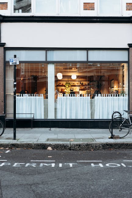 This image shows the exterior of a commercial property with large glass windows revealing a warmly lit interior space, likely a café or restaurant, with white curtains partially covering the windows.